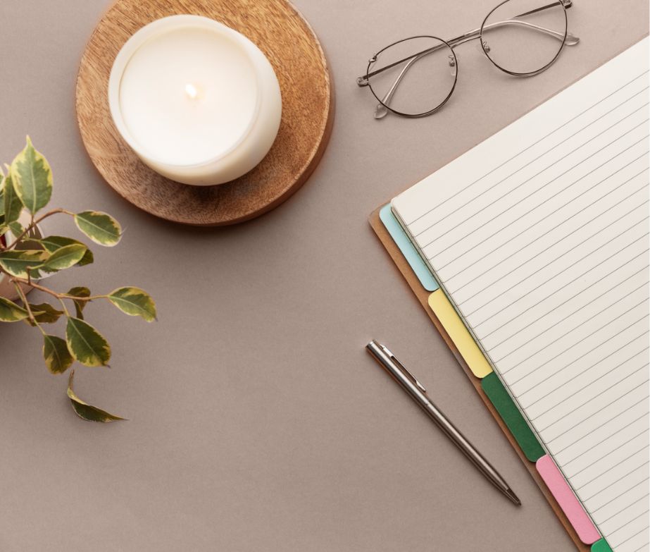 A candle on a wooden coaster, eyeglasses, a leafy plant, a pen, and an open notebook with colorful tab dividers are arranged on a beige surface.