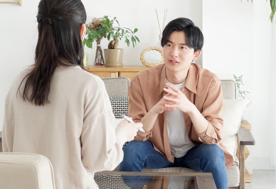 A young man sits on a sofa, talking to a woman holding a notepad. They appear to be having a serious conversation in a bright, cozy living room with plants and wooden decor in the background.
