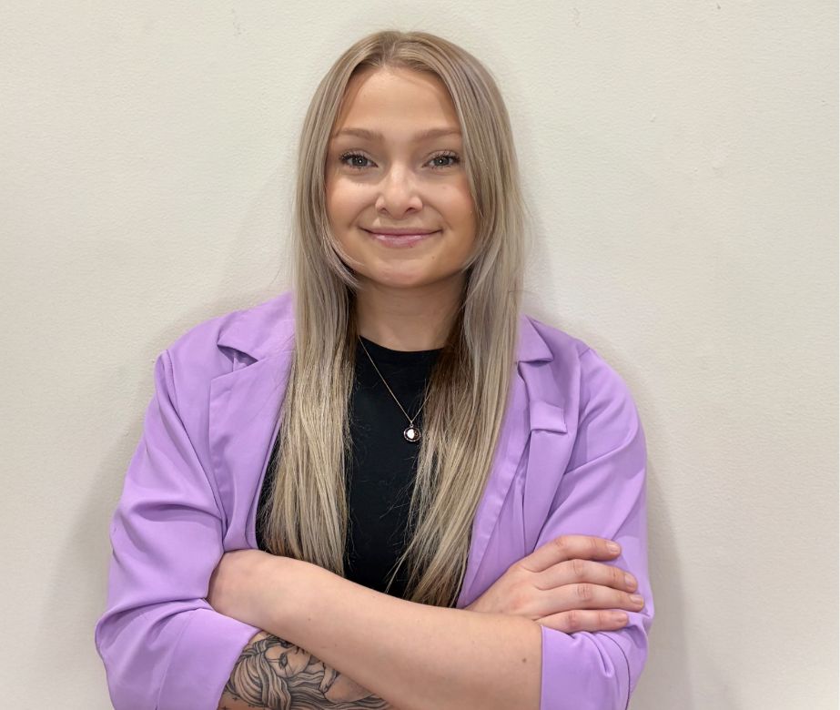 A woman with long, straight blonde hair stands against a plain light-colored wall, smiling with arms crossed. She wears a lavender blazer over a black top and has a floral tattoo on her left forearm.