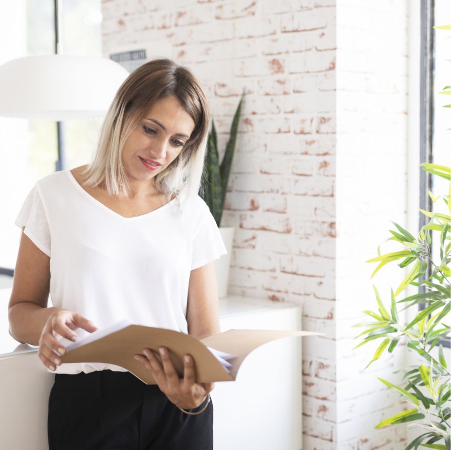 A woman with shoulder-length blonde hair, wearing a white shirt and black pants, stands indoors near a window and plants, looking at papers in a brown folder with a slight smile.
